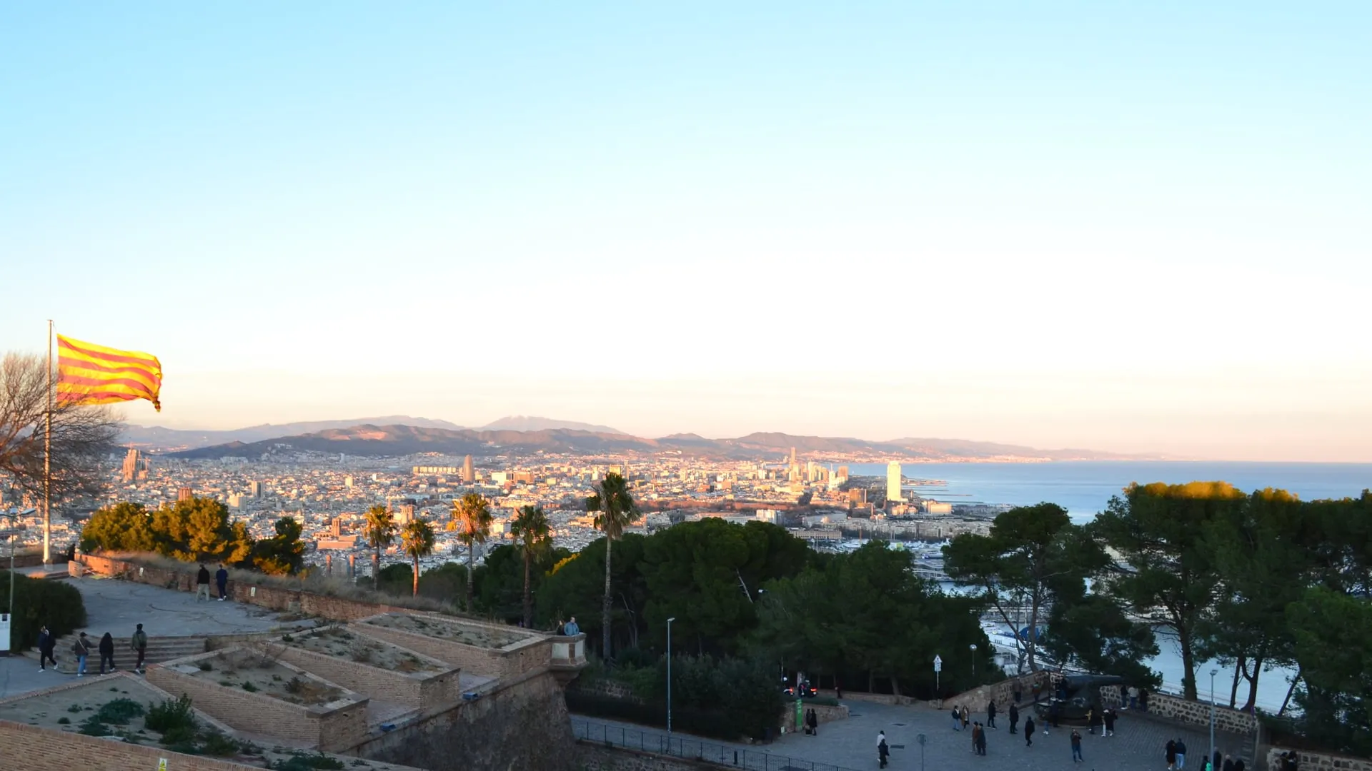Barcelona vista desde lo alto de Montjuïc: la ciudad se extiende hasta el Mediterráneo, las colinas al fondo, una senyera a la izquierda, con una luz de invierno baja y dorada.