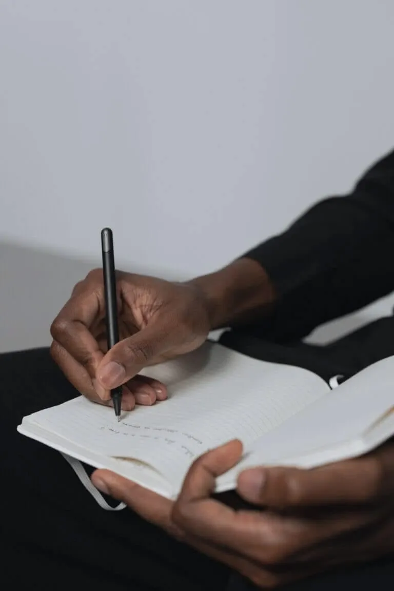 Unas manos oscuras sostienen un cuaderno de rayas abierto y escriben en él con un bolígrafo negro, sobre ropa oscura.